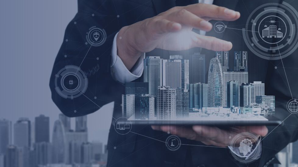 Mani di una persona in abito formale che sorreggono un tablet dal quale emerge una rappresentazione digitale di una città con grattacieli. Intorno appaiono icone trasparenti che simboleggiano tecnologie connesse: AI, edifici intelligenti, Wi-Fi, reti